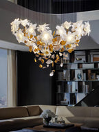 Wide and low over a sofa and tall bookcase, this Clérachic chandelier of white petals and brass leaves is shown in a high-ceilinged lounge.