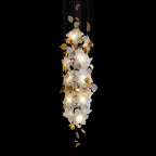 A slim, straight column of lit white petals and brass leaves, this Clérachic chandelier is shown against a black backdrop.