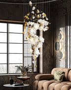 By a tall paned window and a tan sofa, this Clérachic chandelier trails an S of white petals and gold leaves against a rough dark wall.