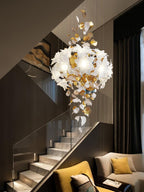 Over a glass-sided staircase and a mustard armchair, this Clérachic chandelier shows a round cluster of petals and trailing brass leaves.
