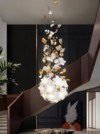 In a stairwell with a curved wooden rail, this Clérachic chandelier hangs in a tall form, brass leaves above a round ball of white petals.