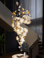 Above a round side table with a book and a vase, this Clérachic chandelier curves down a dark stairwell in lit white petals and brass.