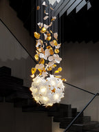 Above a black stair with open treads, this Clérachic chandelier hangs with brass leaves on top and a round lit ball of white petals below.