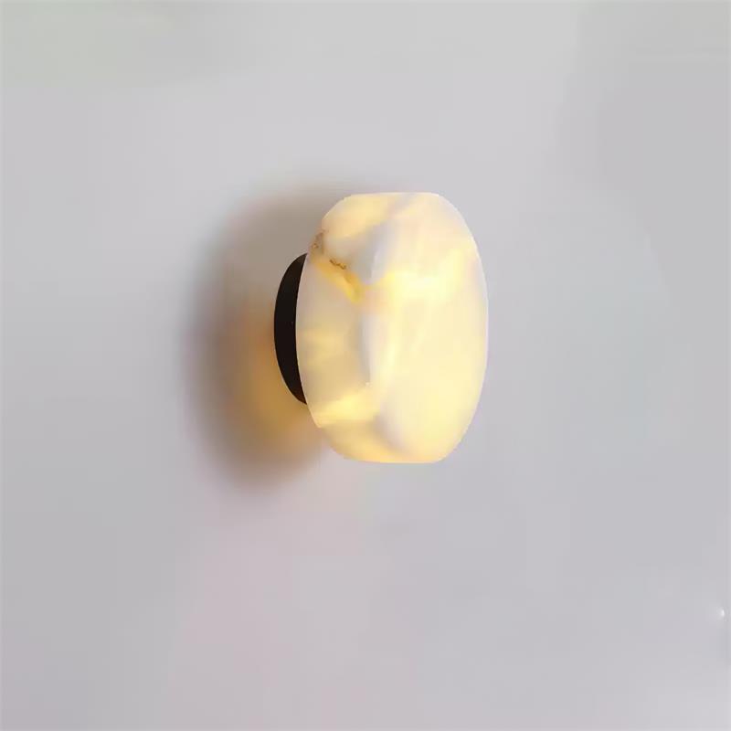The black-backplate Clérachic Glim Round Alabaster Wall Sconce seen at an angle, warm light pooling on the wall behind the stone.