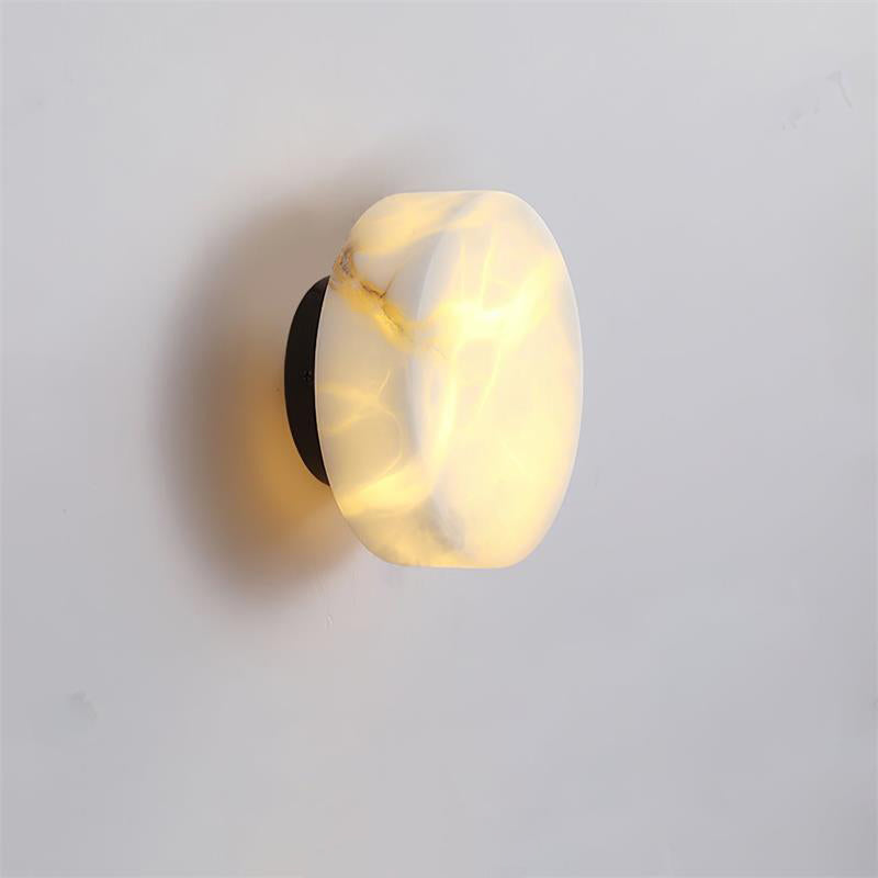 Lit Clérachic Glim Round Alabaster Wall Sconce on a black backplate, the stone face reading as a soft warm disc in a quiet room.