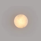 Face-on view of a smaller Clérachic Glim Round Alabaster Wall Sconce, its lit stone reading as a plain warm circle on a grey wall.