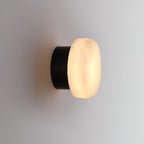 Side view of the Clérachic Glim Round Alabaster Wall Sconce, the lit stone face sitting over a short black cylindrical backplate.