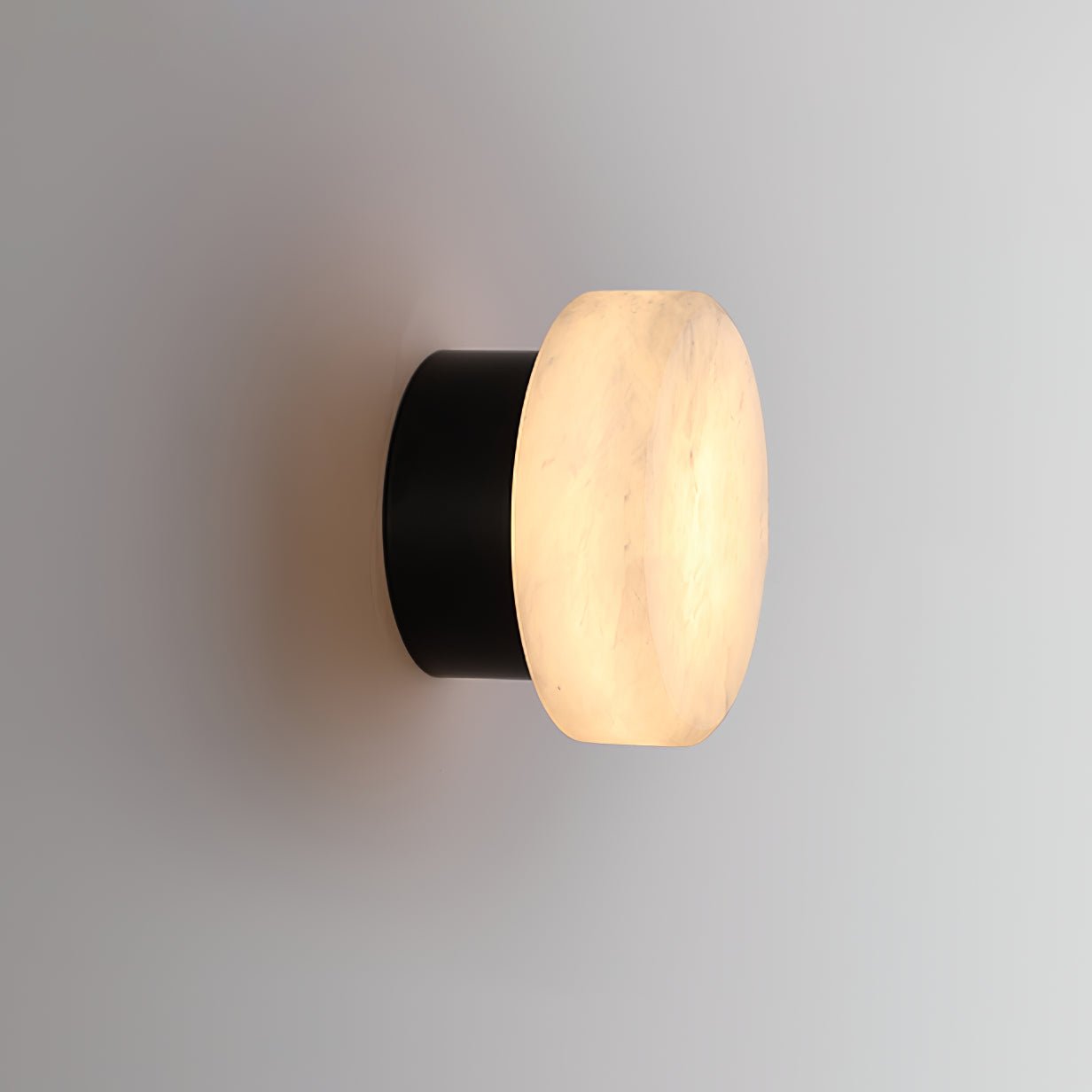 Side view of the Clérachic Glim Round Alabaster Wall Sconce, the lit stone face sitting over a short black cylindrical backplate.