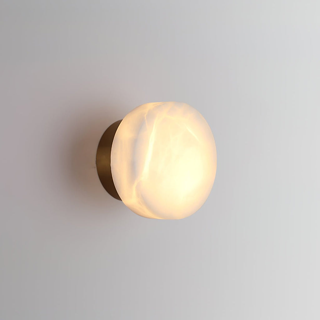 Brass-backed Clérachic Glim Round Alabaster Wall Sconce lit at an angle, the glow washing down the wall beneath the stone.