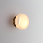 Brass-backed Clérachic Glim Round Alabaster Wall Sconce lit at an angle, the glow washing down the wall beneath the stone.