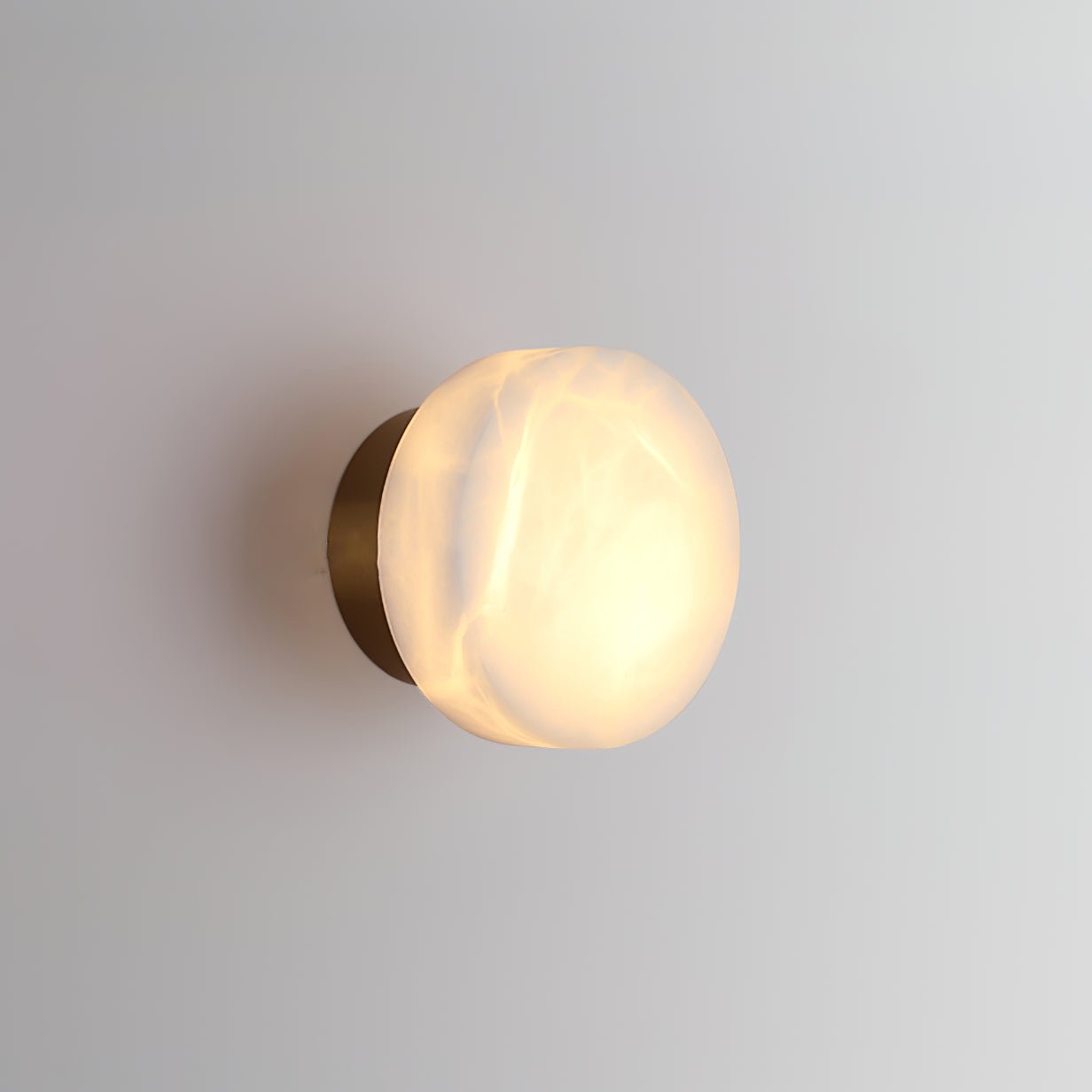 Brass-backed Clérachic Glim Round Alabaster Wall Sconce lit at an angle, the glow washing down the wall beneath the stone.