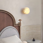 A lit Clérachic Glim Round Alabaster Wall Sconce above an arched wooden headboard, reading as a soft disc over a travertine bedhead.