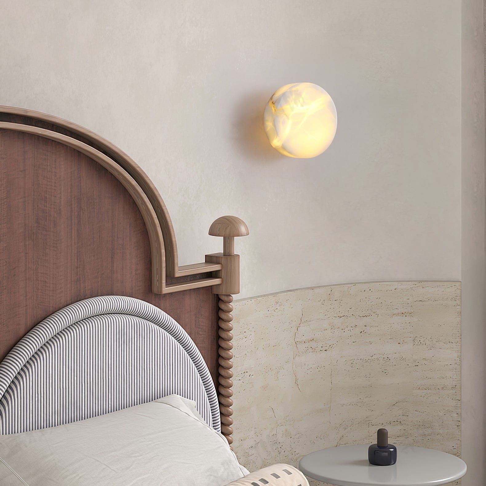 A lit Clérachic Glim Round Alabaster Wall Sconce above an arched wooden headboard, reading as a soft disc over a travertine bedhead.