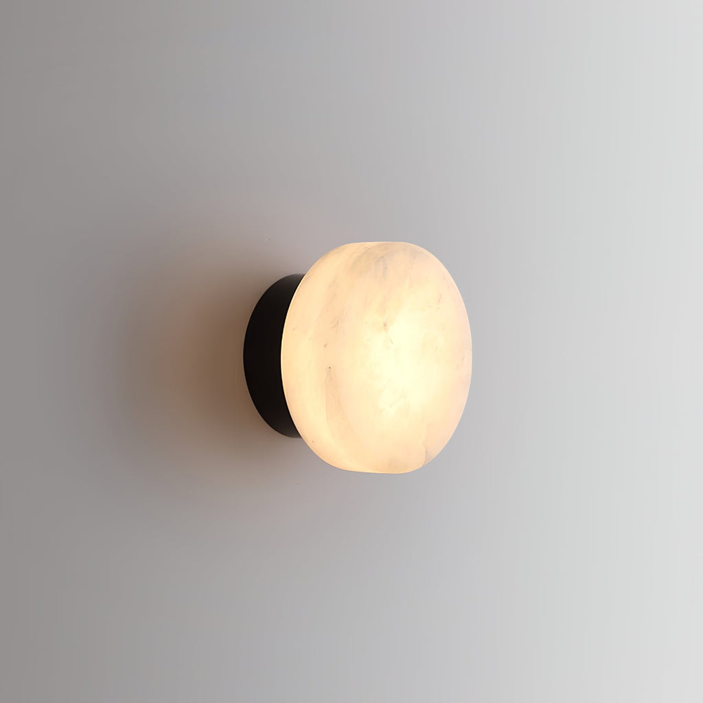 The Clérachic Glim Round Alabaster Wall Sconce in black, lit softly so the stone's faint markings show without any hot spot.