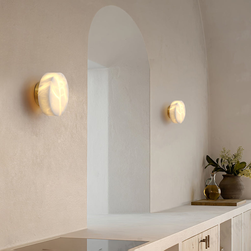Two brass-backed Clérachic Glim Round Alabaster Wall Sconces lit either side of an arched doorway in a pale limewash kitchen.
