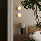 Two Clérachic Glim Round Alabaster Wall Sconces stacked and lit on a bedroom wall beside a rattan bed and a bedside lamp.