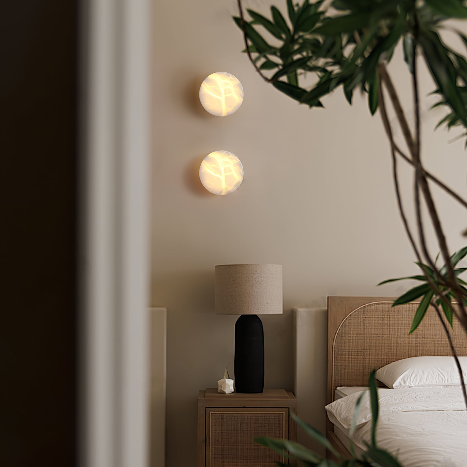 Two Clérachic Glim Round Alabaster Wall Sconces stacked and lit on a bedroom wall beside a rattan bed and a bedside lamp.