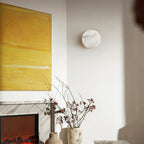 An unlit Clérachic Glim Round Alabaster Wall Sconce on a white living room wall, beside a marble fireplace and a yellow canvas.