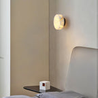 A lit Clérachic Glim Round Alabaster Wall Sconce in black on a bedroom corner wall, above a slim black bedside shelf.