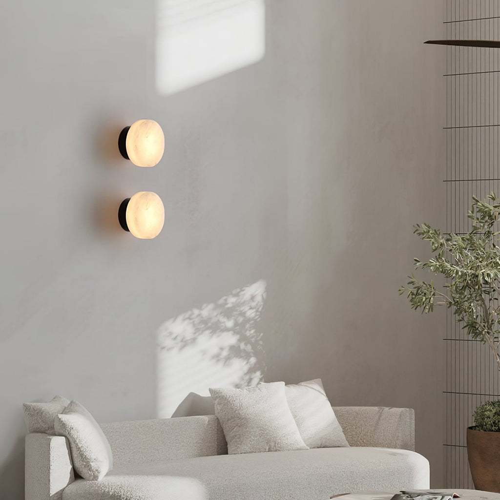Two black-backed Clérachic Glim Round Alabaster Wall Sconces lit one above the other on a limewash wall behind a pale sofa.