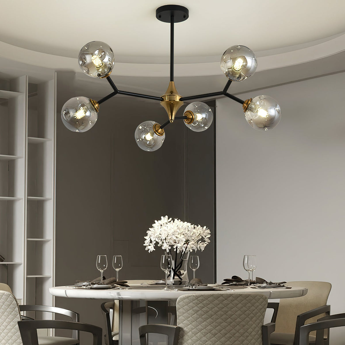 A smaller Clérachic chandelier with grey glass globes hangs over a round marble dining table set with glasses and white flowers.