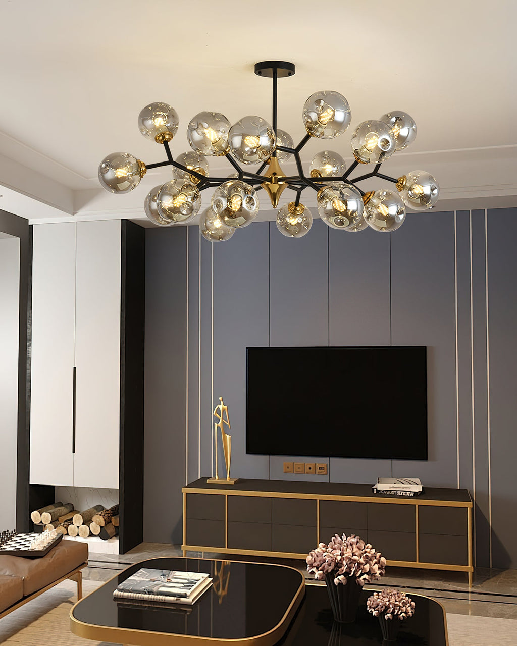 A Clérachic chandelier with smoky grey globes hangs over a living room with a wall-mounted television and a gold-edged cabinet.