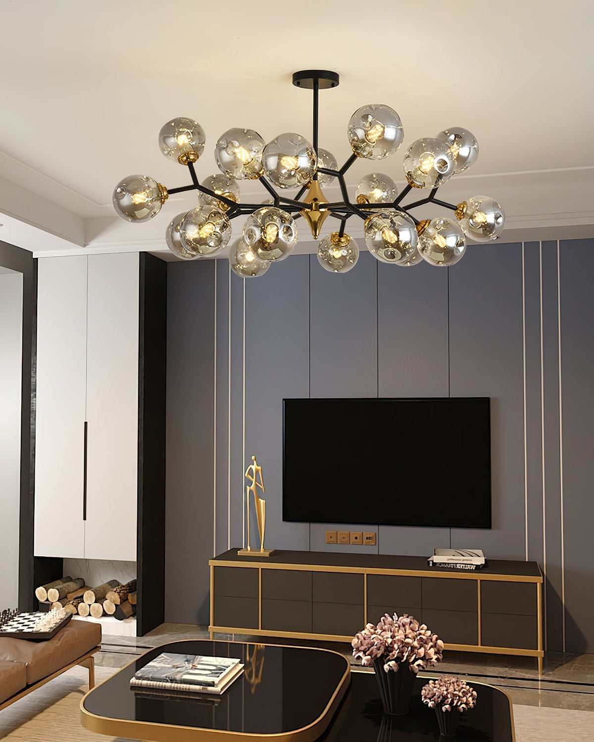 A Clérachic chandelier with smoky grey globes hangs over a living room with a wall-mounted television and a gold-edged cabinet.