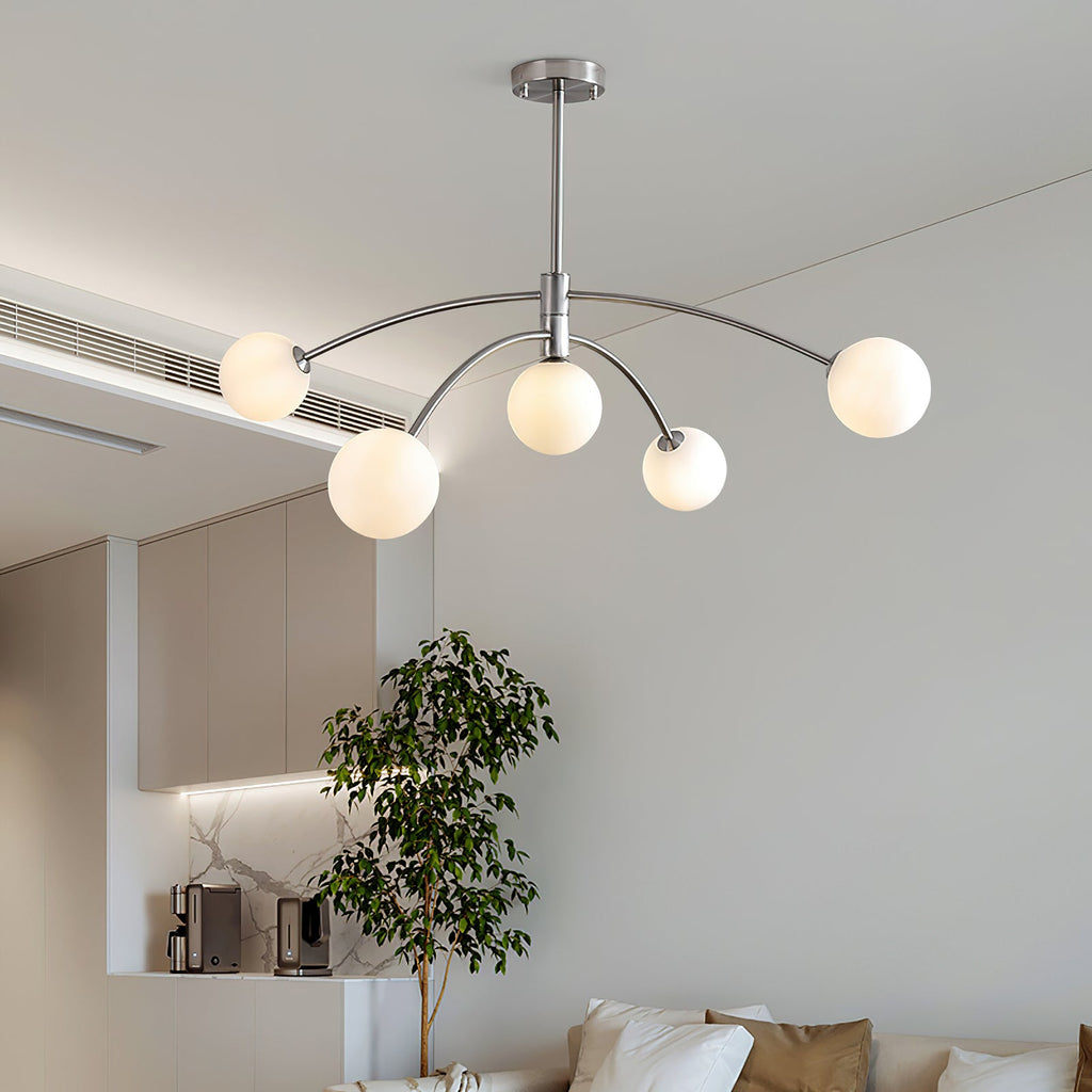 Chrome-finish Clérachic chandelier with five frosted globes on curved arms, hanging in a pale room with a tall potted tree beyond.