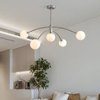 Chrome-finish Clérachic chandelier with five frosted globes on curved arms, hanging in a pale room with a tall potted tree beyond.