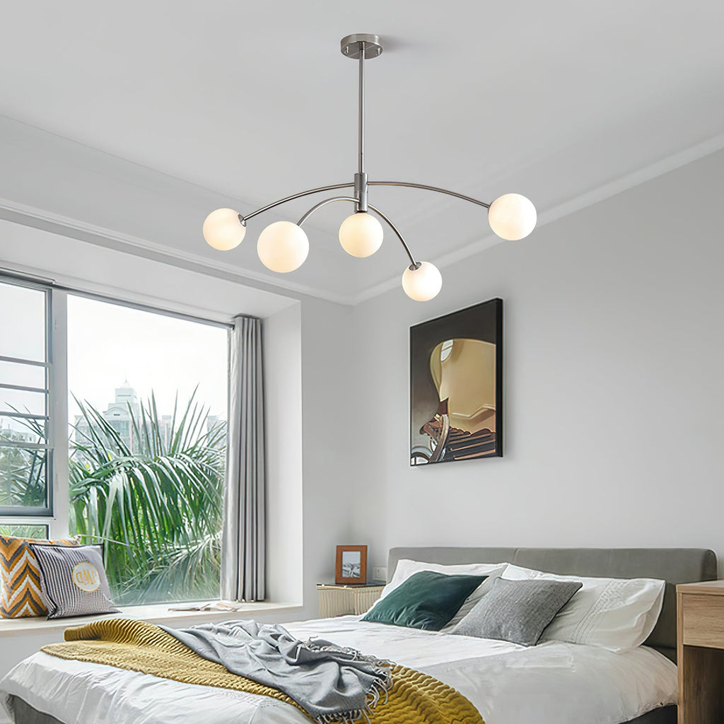 Chrome Clérachic globe chandelier above a bed in a bright bedroom with a yellow throw, green cushions and a palm outside the window.