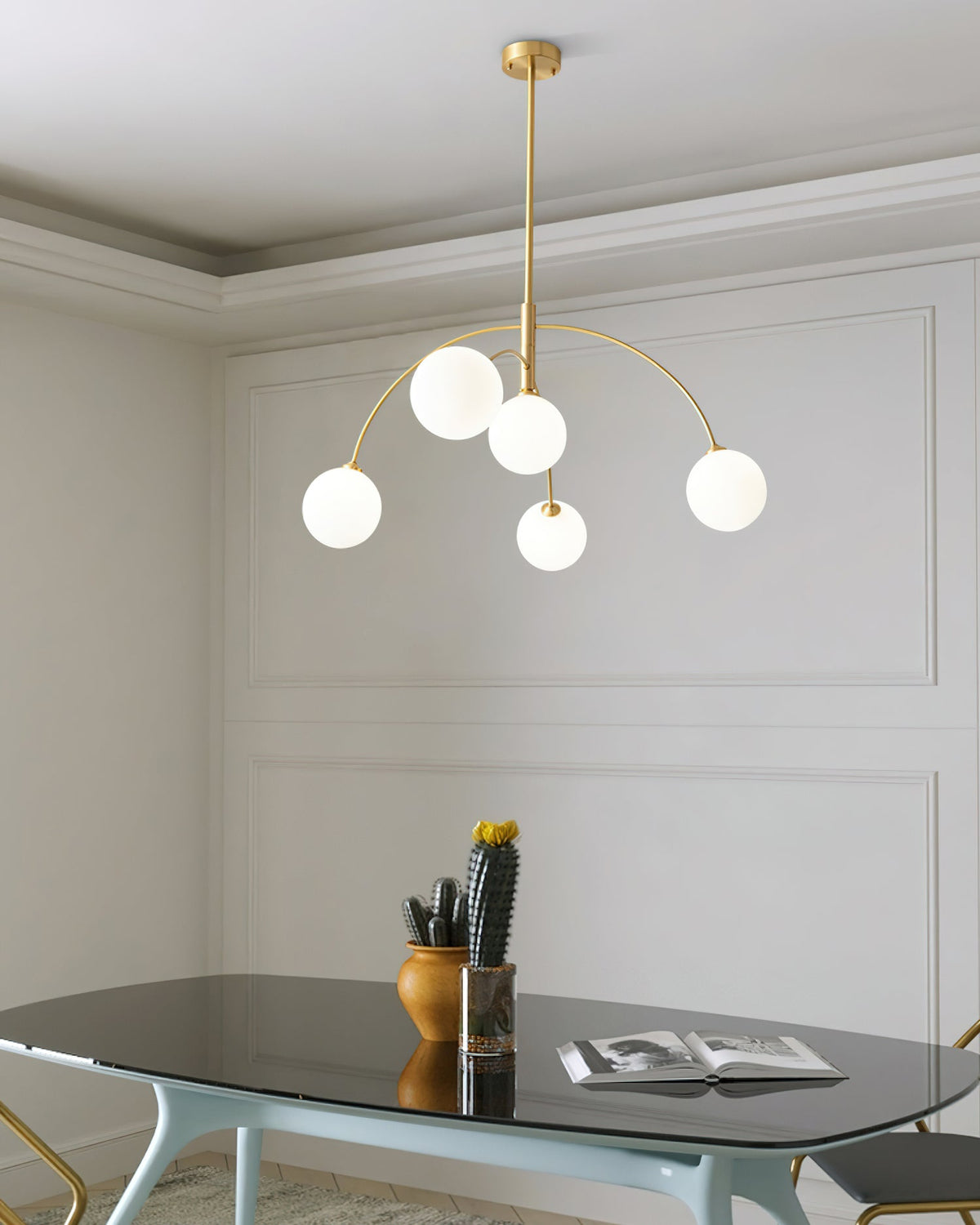 Gold-finish Clérachic chandelier with five lit globes in a panelled room, above a glass-topped table with a cactus and an open book.