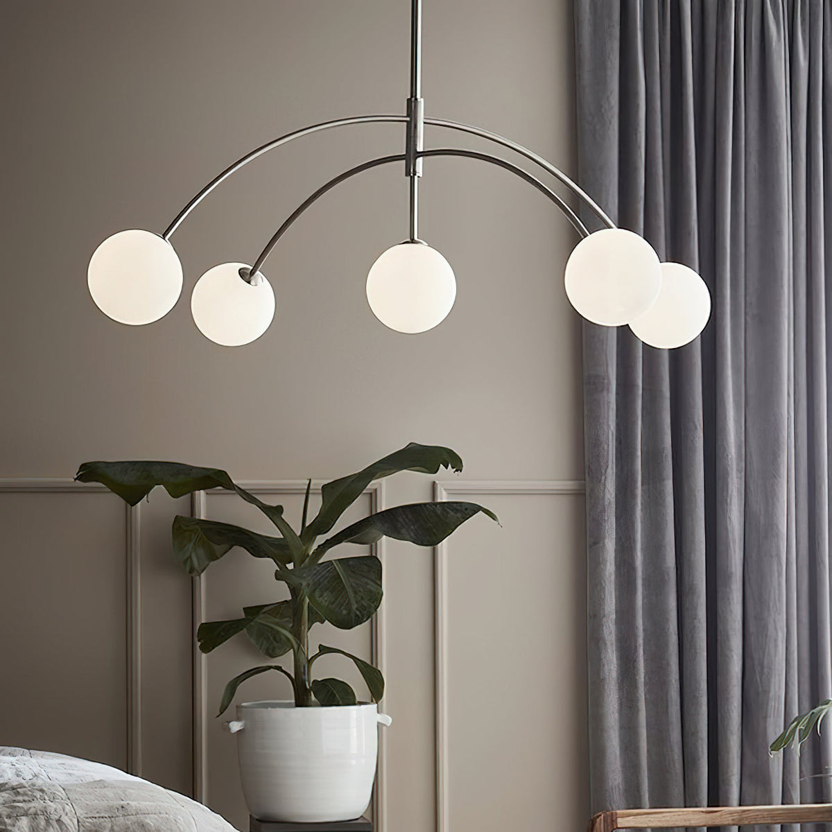 Chrome Clérachic chandelier above a grey room with heavy curtains and a large leafy plant, its globes at different heights.
