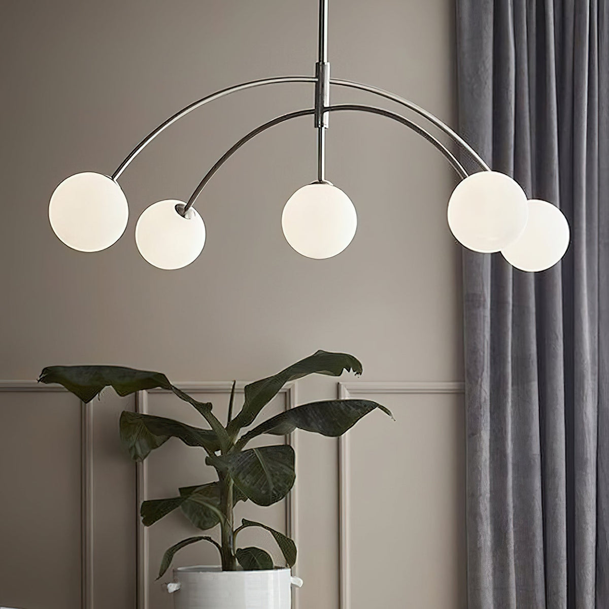 Close view of the chrome Clérachic chandelier's arched arms and frosted globes, with grey curtains and a leafy plant behind.