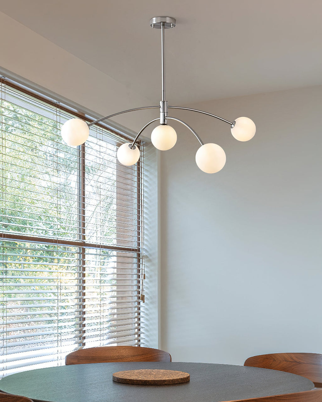 Chrome Clérachic chandelier lit above a round dining table, with a window of slatted blinds behind it.