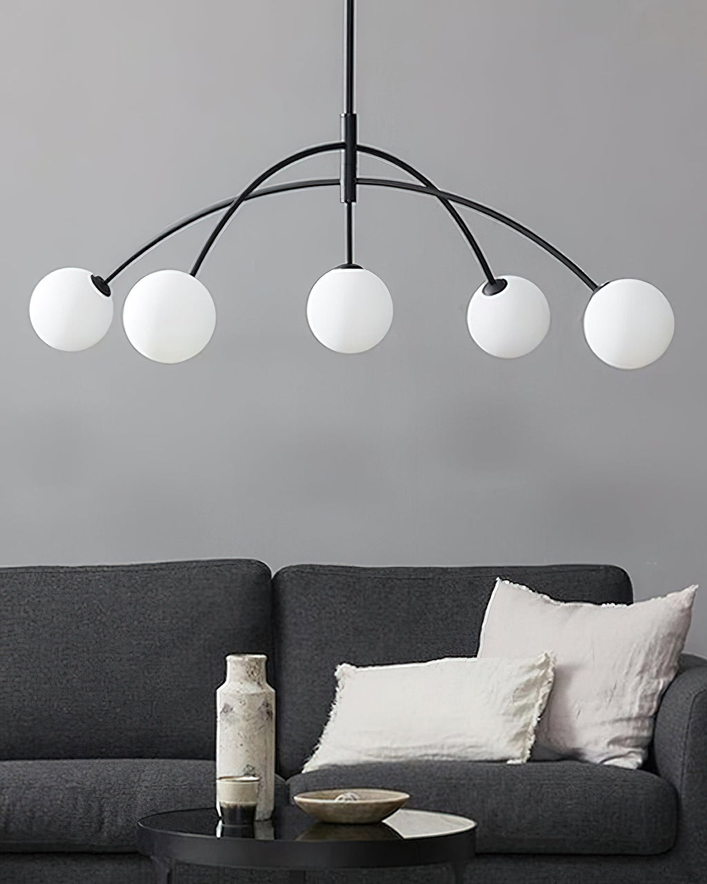 Black and white Clérachic chandelier with five frosted globes, hanging in front of a grey wall above a dark sofa.