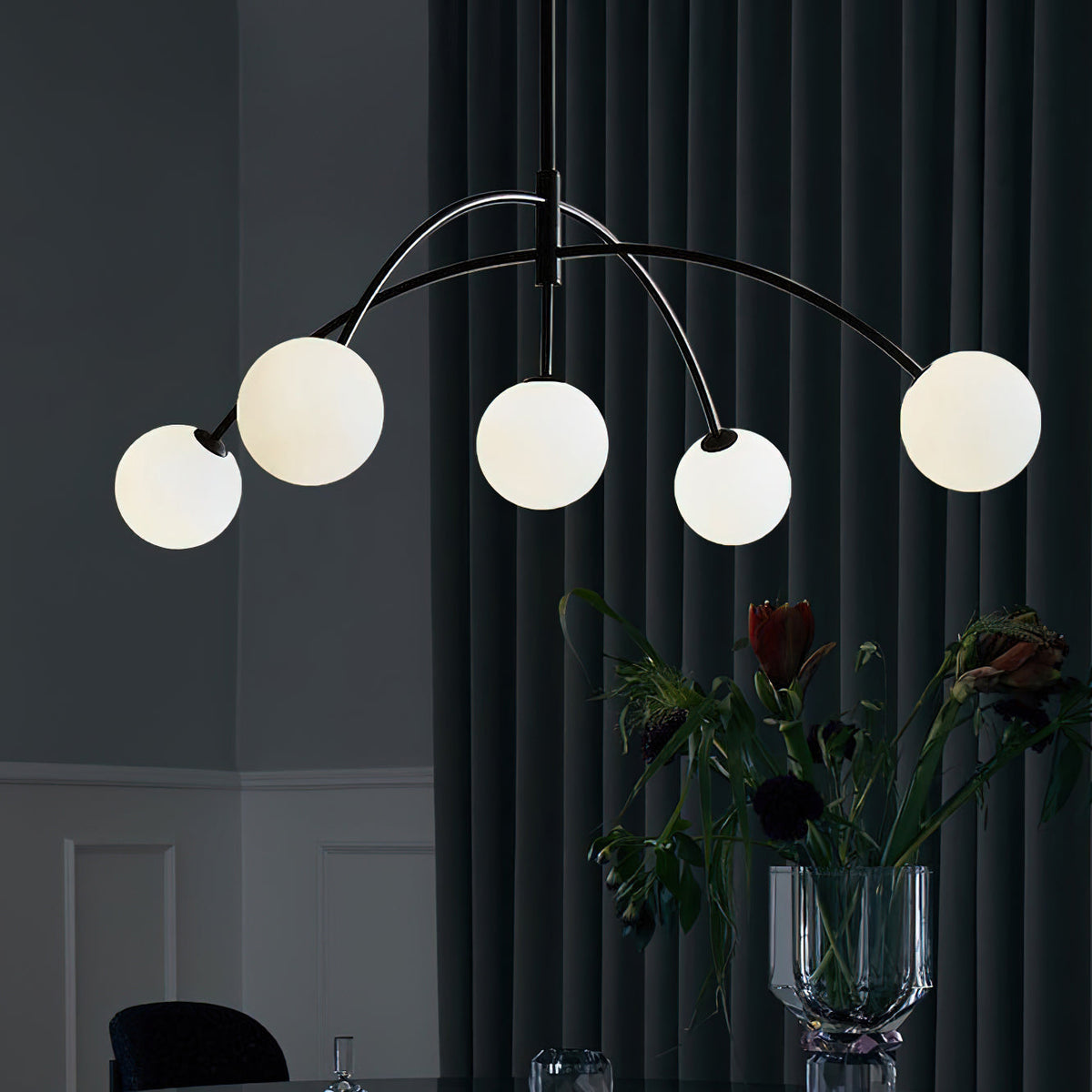 Black and white Clérachic chandelier against a dark curtain, its globes lit and a vase of flowers on the table below.