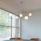 Chrome Clérachic chandelier in a bright room with slatted blinds and a view of trees, above a dining table.