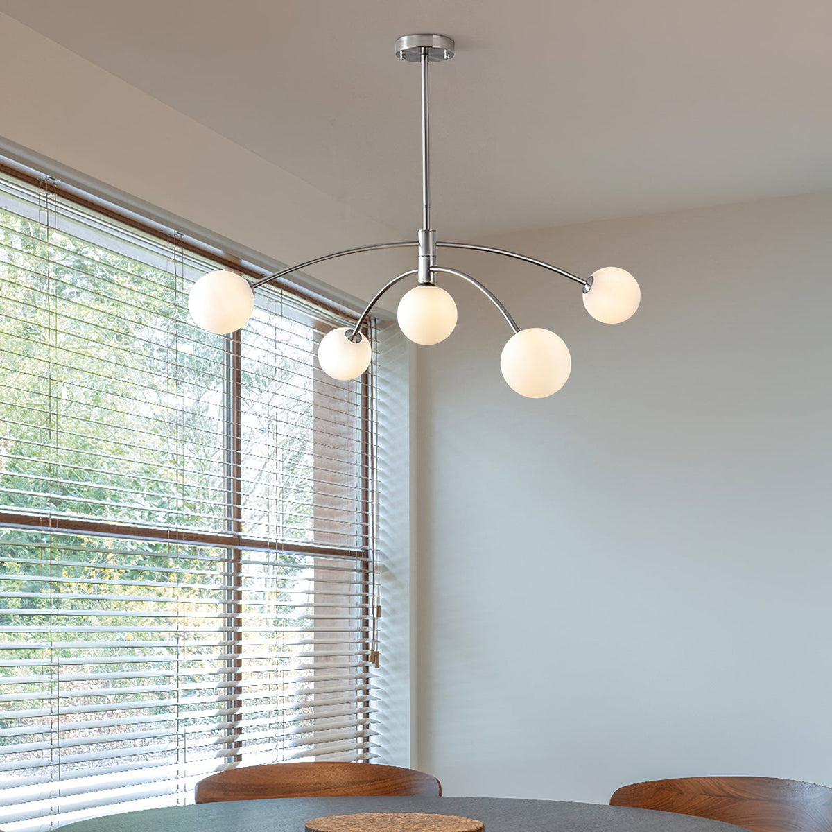 Chrome Clérachic chandelier in a bright room with slatted blinds and a view of trees, above a dining table.