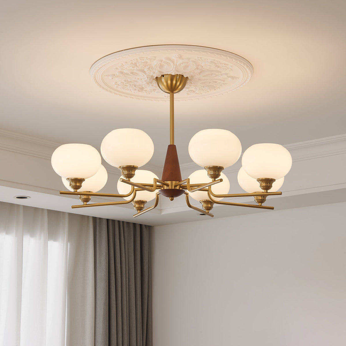 Under a carved plaster ceiling rose, a Clérachic chandelier with milky globes and brass arms hangs beside grey and white curtains.