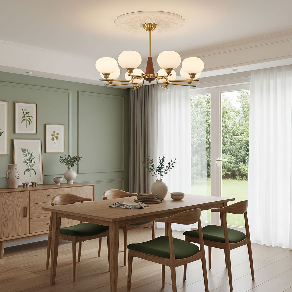 Over a pale wood dining table with green seat pads, a Clérachic chandelier hangs in a sage-green panelled room with garden doors.