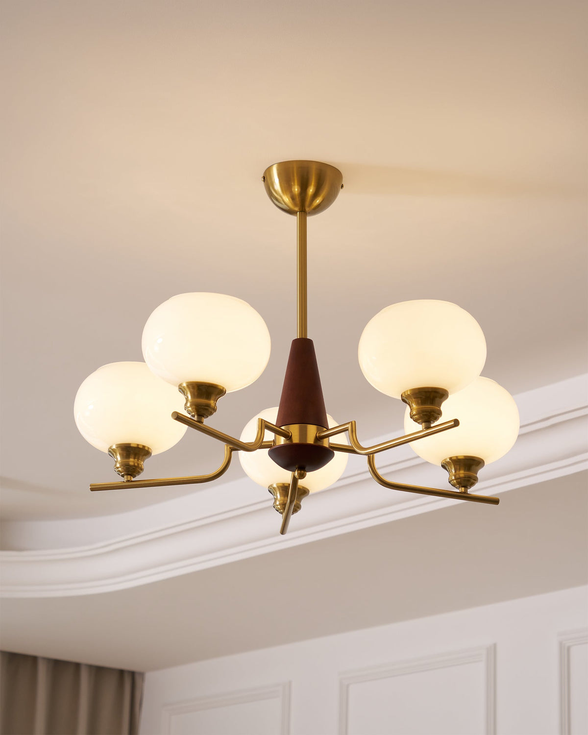 Close view of a Clérachic chandelier, with a brass canopy and rod, a walnut cone and glowing frosted globes, by a moulded cornice.