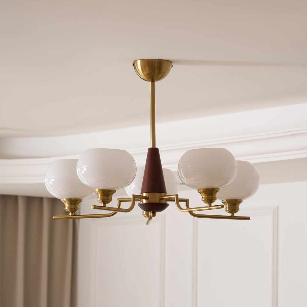 Looking up, this Clérachic chandelier shows rounded white globes on slim brass arms, with grey curtains at the window behind.