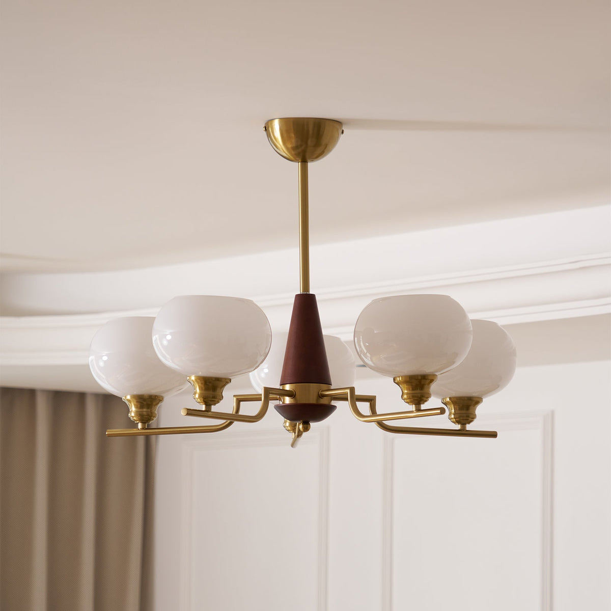 Looking up, this Clérachic chandelier shows rounded white globes on slim brass arms, with grey curtains at the window behind.