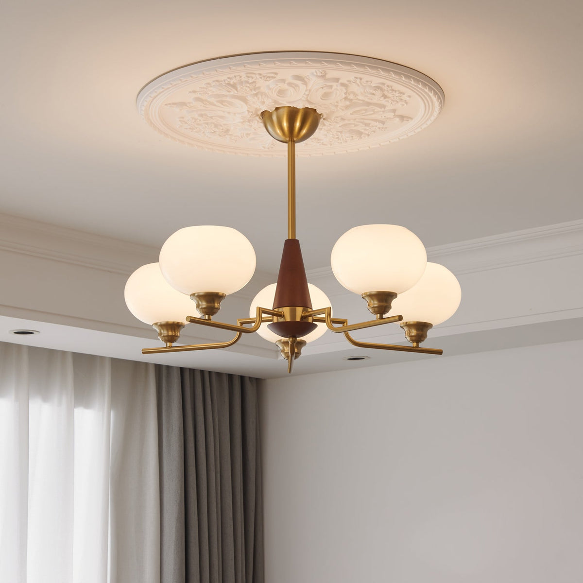 The milky globes of this Clérachic chandelier glow beneath an ornate plaster ceiling rose, beside curtains at the window.