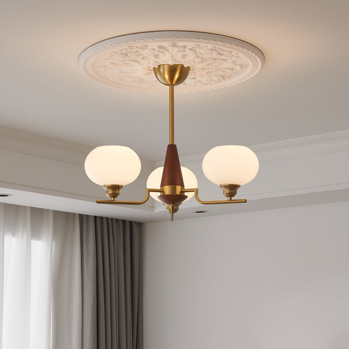 A three-light Clérachic chandelier with a brass rod and walnut cone hangs below a plaster ceiling rose, beside soft curtains.