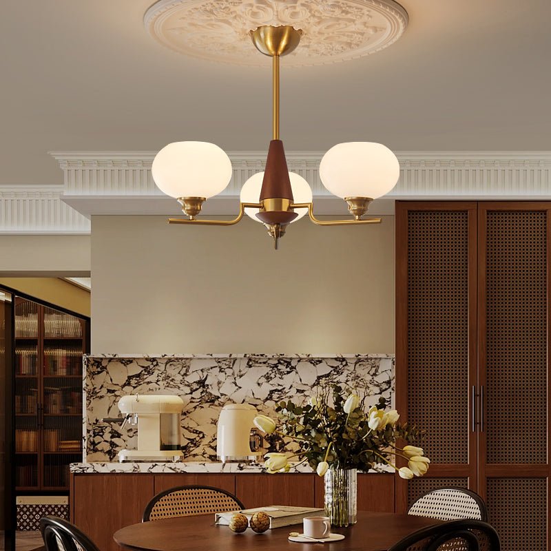 A Clérachic chandelier with three globes hangs over a dining area with round cane-back chairs and a marble splashback.