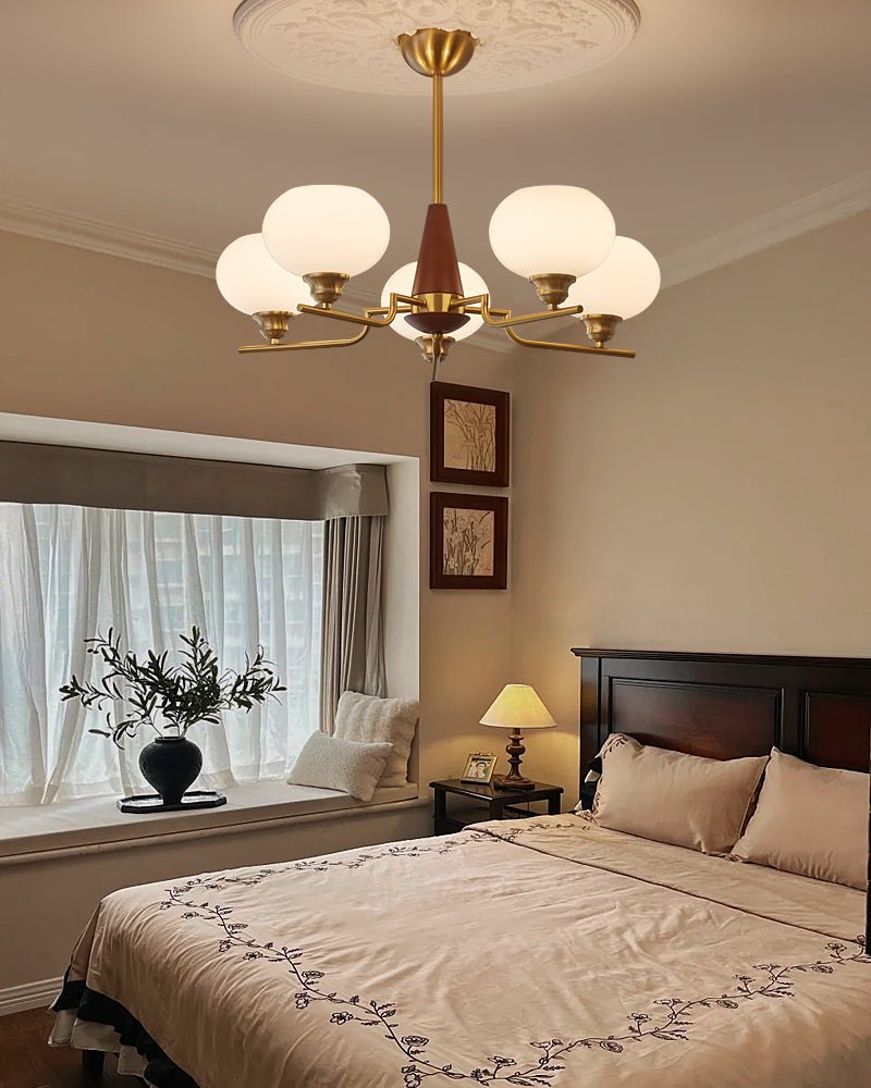 The frosted globes and brass arms of a Clérachic chandelier hang over a bed with embroidered bedding, by a bay window.