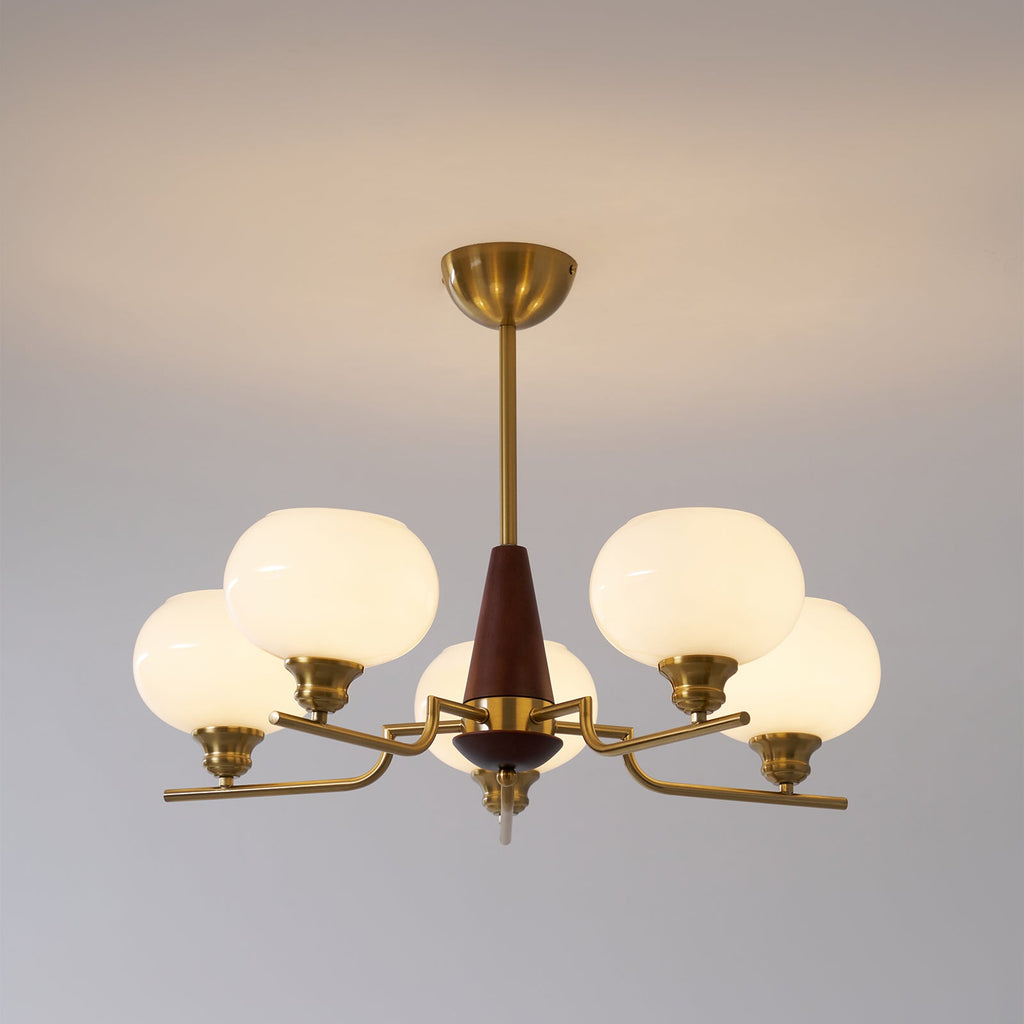 A lit Clérachic chandelier with five frosted globes hangs from a brass rod against a soft grey wall.