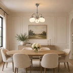 Above an oval wooden dining table with cream chairs, a Clérachic chandelier with glowing white globes hangs under a landscape painting.