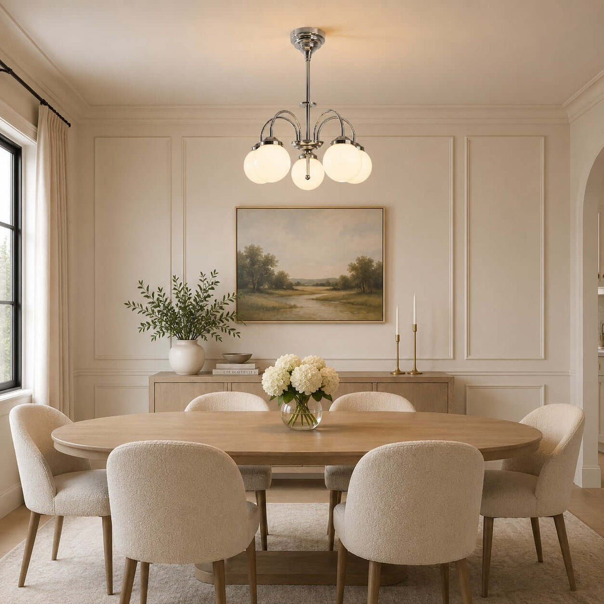 Above an oval wooden dining table with cream chairs, a Clérachic chandelier with glowing white globes hangs under a landscape painting.